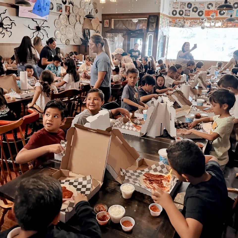 Children and adults gather around tables, making pizzas in a lively restaurant setting.