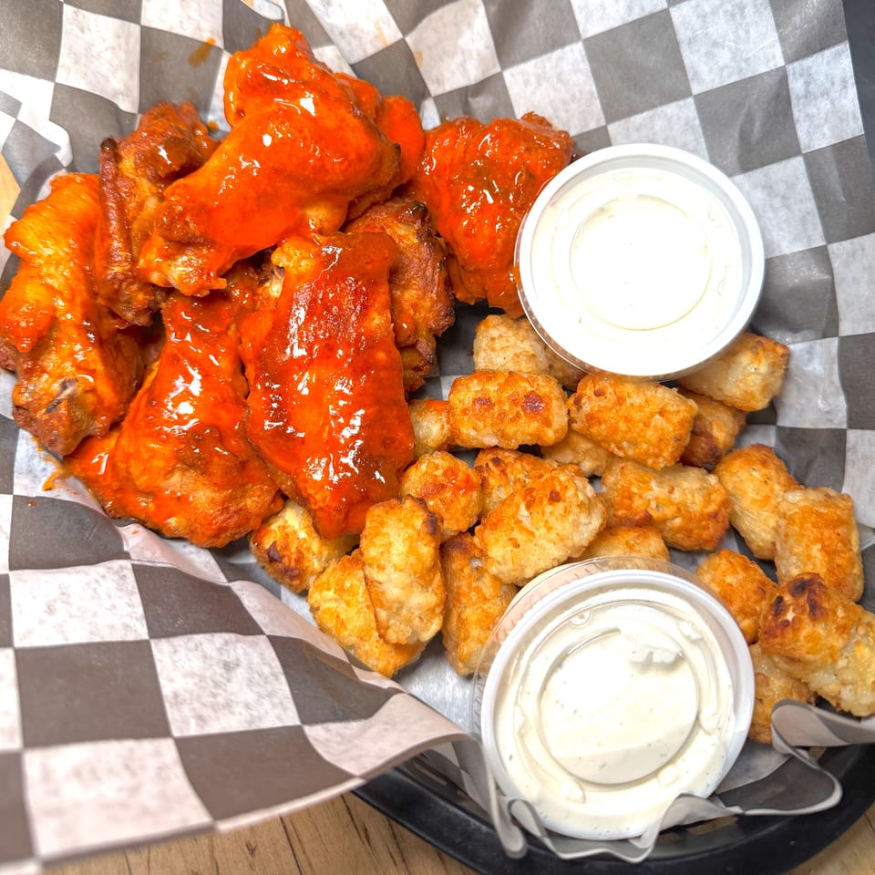 Buffalo wings and tater tots in a basket with two dipping sauces on a checkered paper liner.