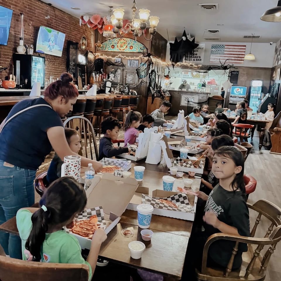 Kids sit at long tables, eating pizza in a restaurant decorated for Halloween, with an adult supervising.