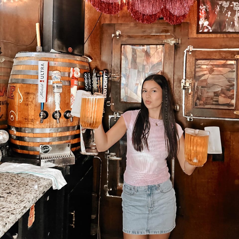 A young woman holds two large pitchers of beer next to a wooden barrel tap system.