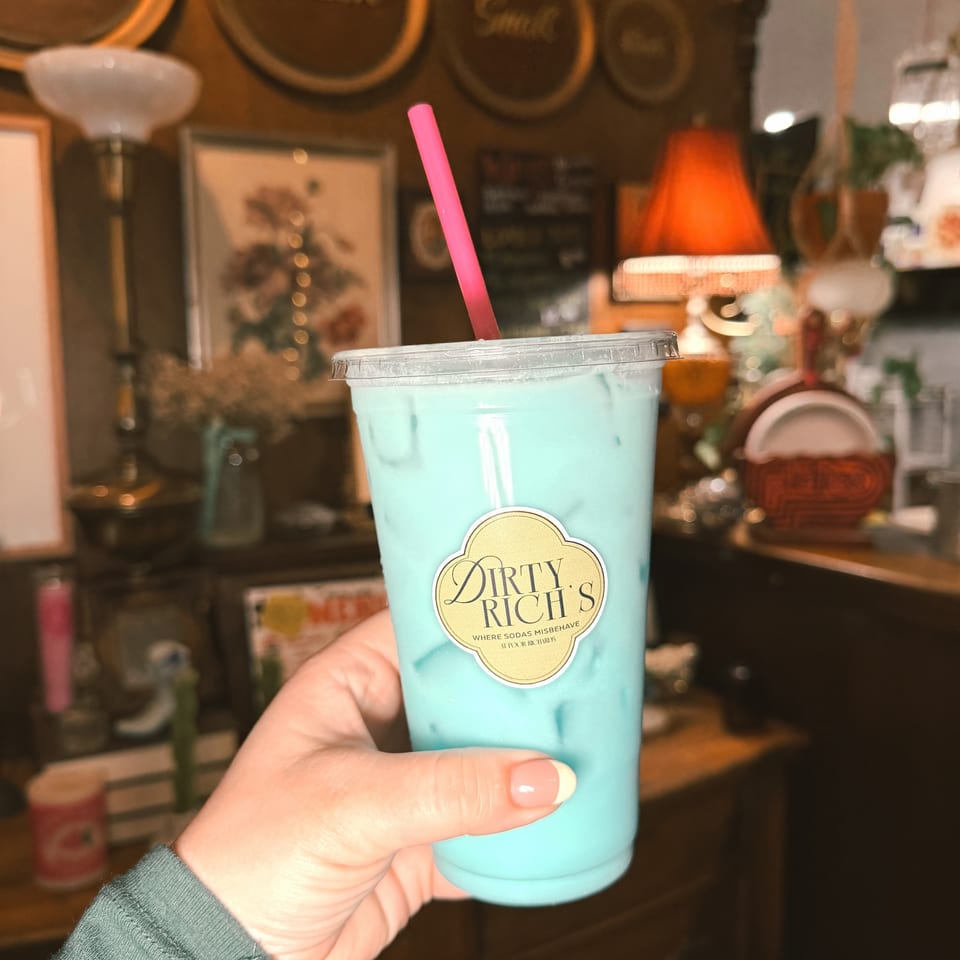 Hand holding a light blue drink with a pink straw and "Dirty Rich's" logo in a cozy, vintage-style room.