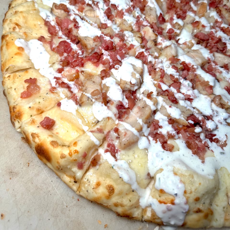 Close-up of a pizza with white sauce, chicken, and bacon bits, cut into squares.