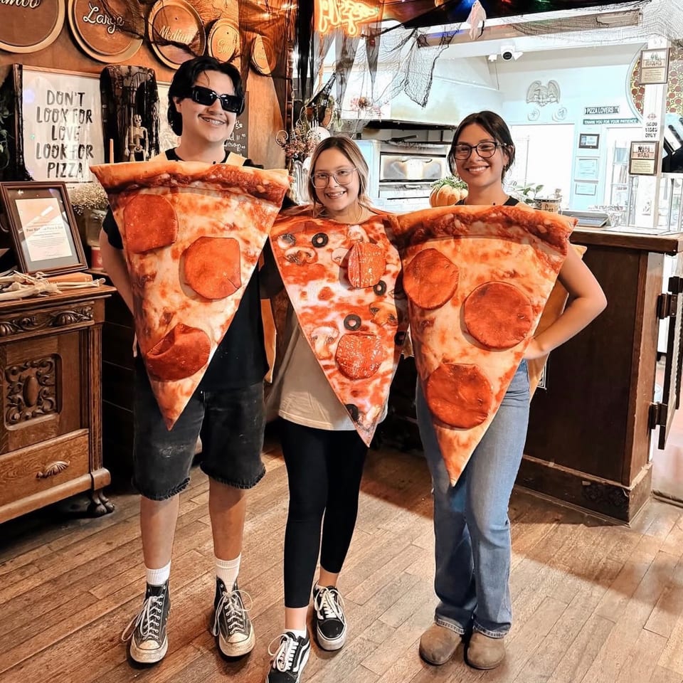 Three young people in pizza slice costumes, two pepperoni and one mushroom and olive, smiling in a restaurant.