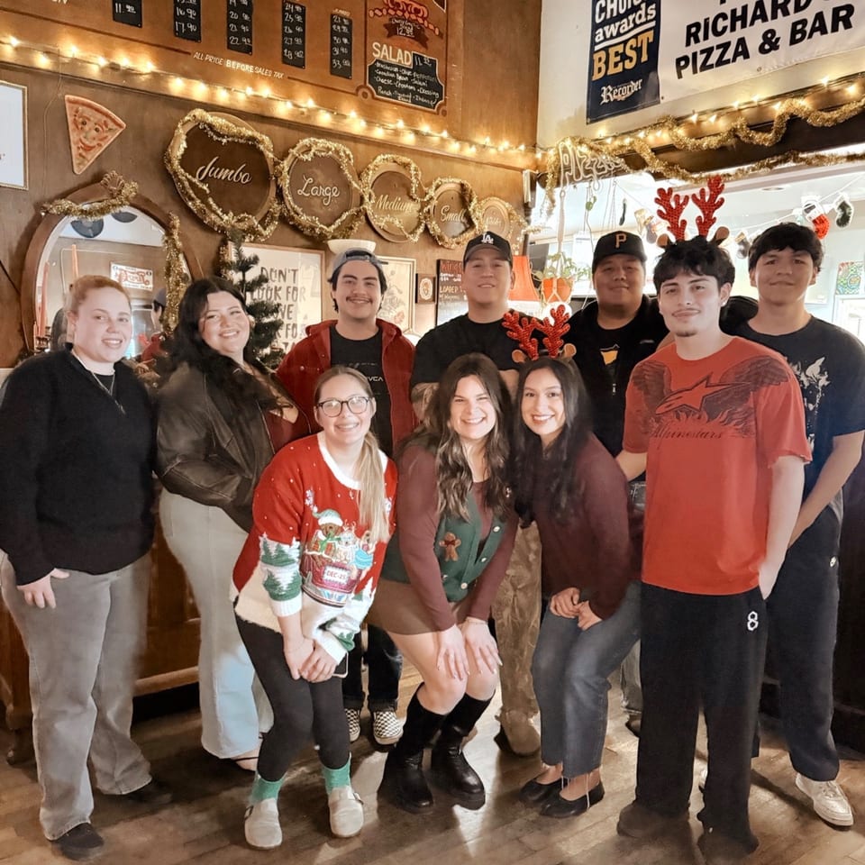 Nine smiling young adults, some in festive attire, pose in a warmly lit pizza restaurant decorated with string lights.