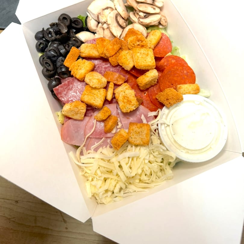 A large chef salad in a white takeout box, with ham, pepperoni, olives, mushrooms, croutons, and cheese.
