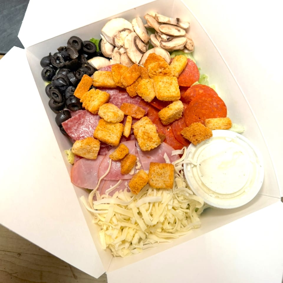A white takeout box filled with a salad of olives, mushrooms, pepperoni, ham, shredded cheese, and croutons.