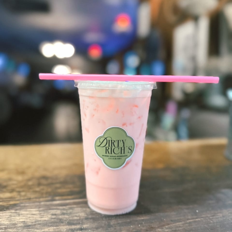 A Dirty Rich's pink drink with ice and a pink straw sits on a wooden surface.
