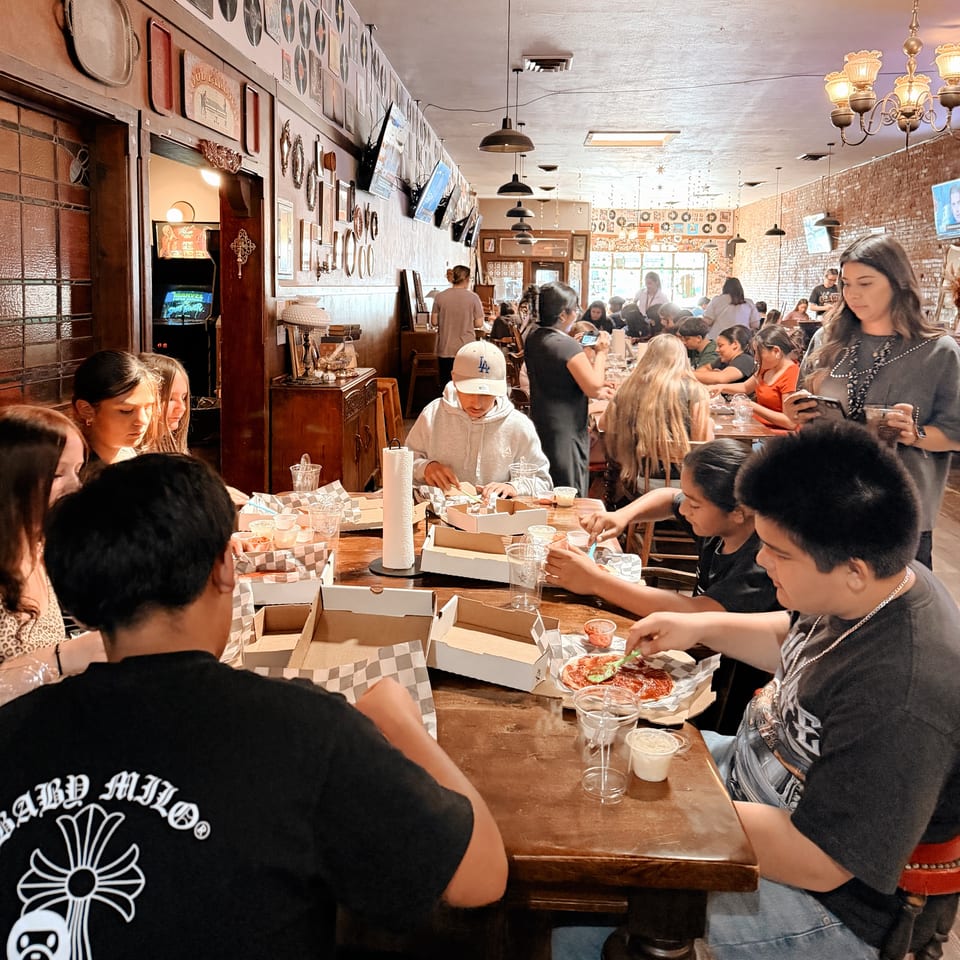 Diners enjoying food at long wooden tables in a busy restaurant with vintage decor and brick walls.