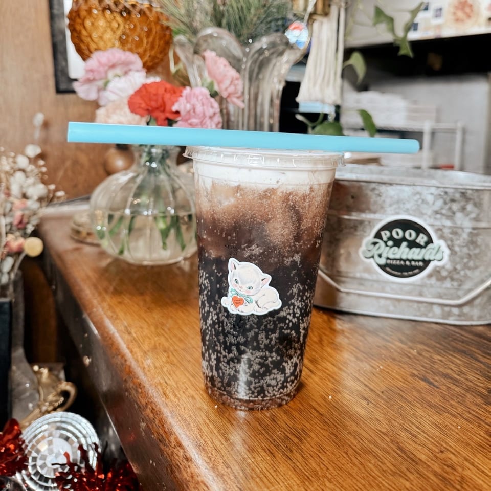 Bubbly dark soda with a white foam top and a cute cat sticker, on a wooden counter.
