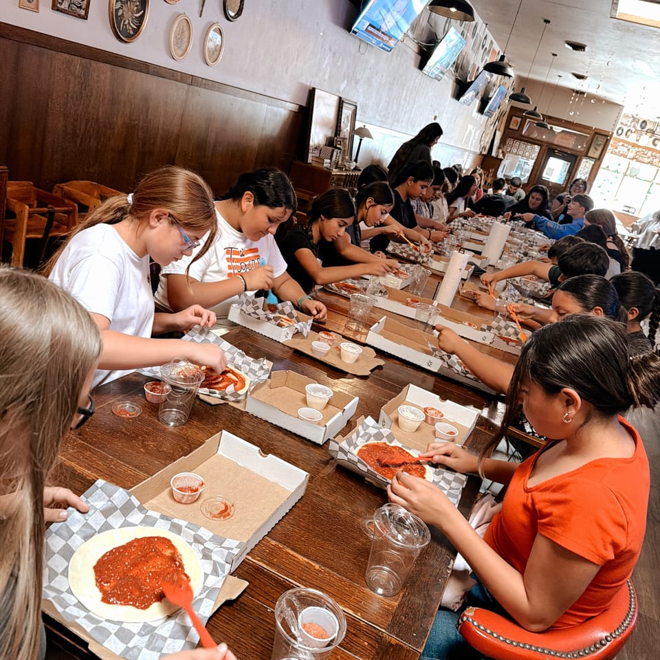 Children at a long wooden table making pizzas with sauce and toppings in a restaurant.