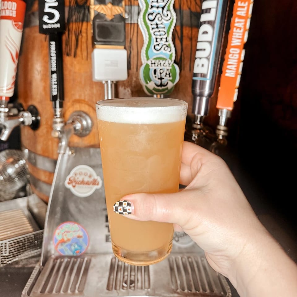 Hand holding a glass of beer with a foamy head, in front of beer taps.