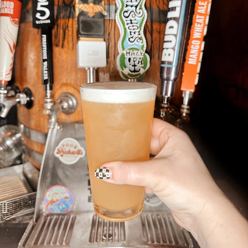 Hand holding a glass of beer with a foamy head, in front of beer taps.