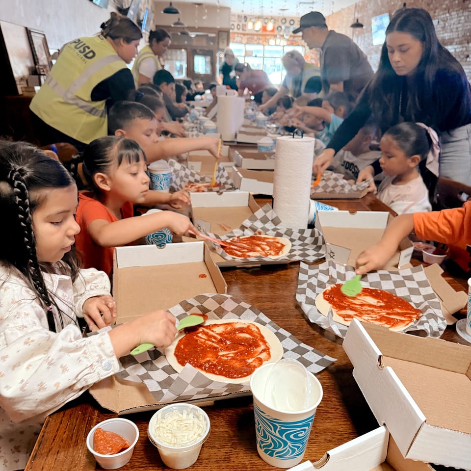 Children and adults making pizzas at a long wooden table, spreading sauce on dough in cardboard boxes.
