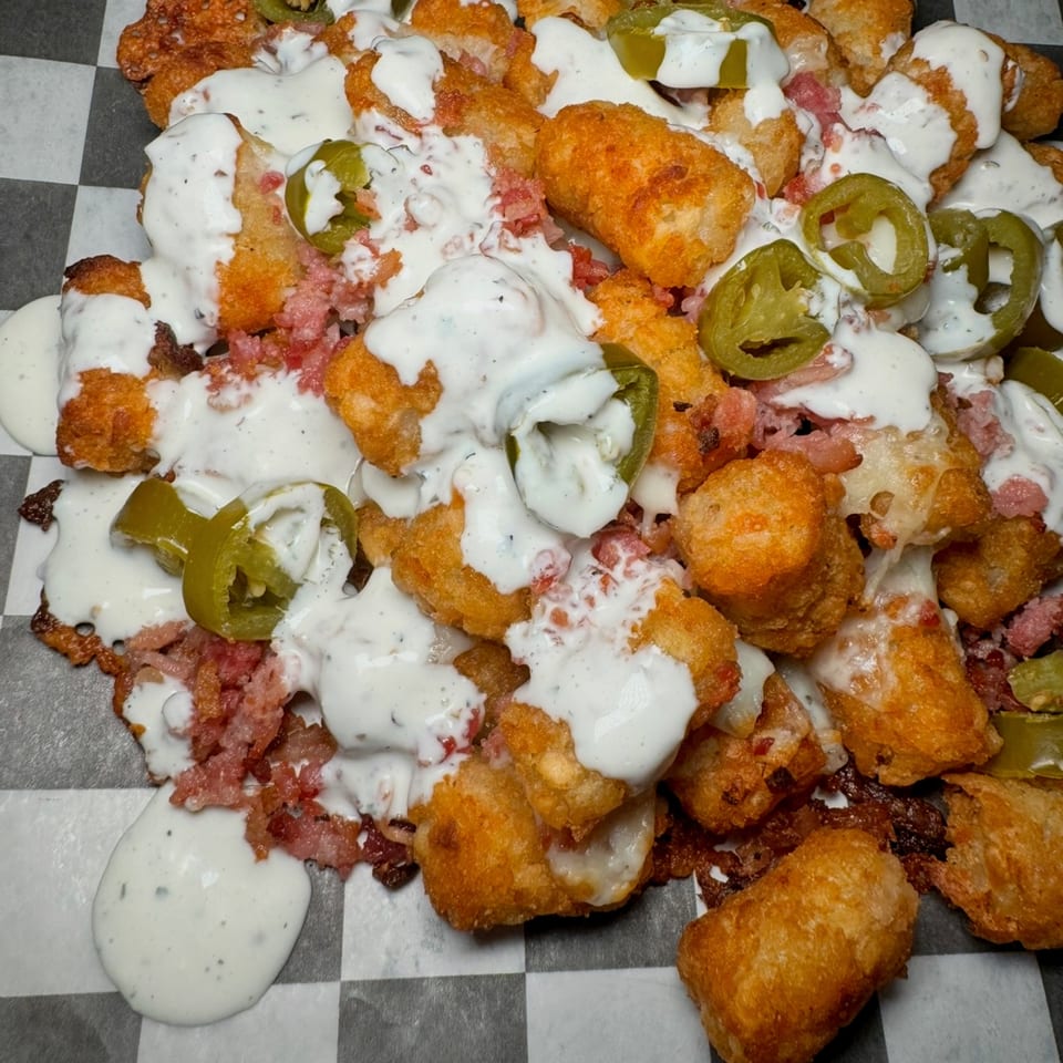 Loaded tater tots with melted cheese, bacon bits, jalapeños, and creamy white sauce on checkered paper.