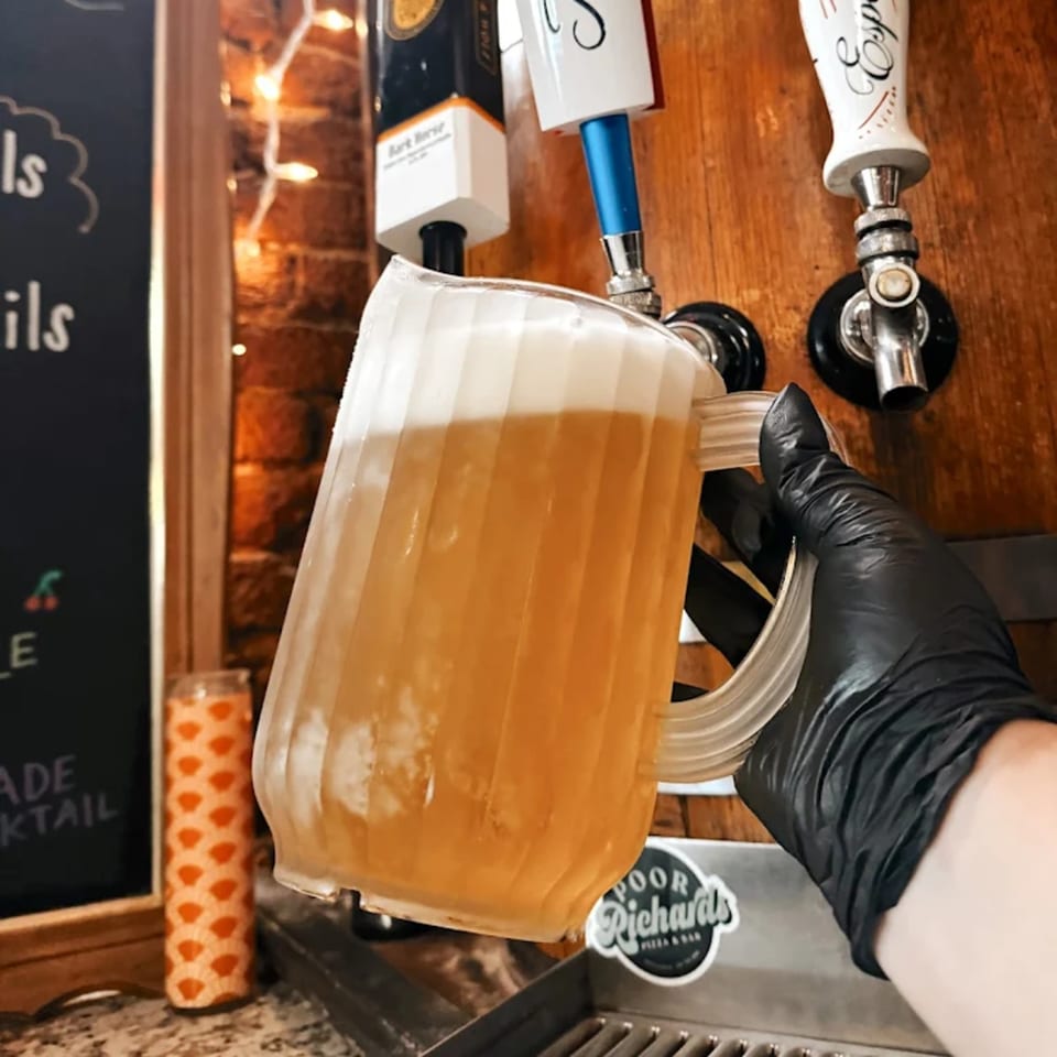 A gloved hand fills a pitcher with a foamy, golden beverage from a tap at Poor Richard's Pizza & Bar.