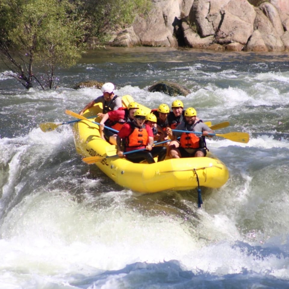 Six people in a yellow raft navigate turbulent whitewater rapids on a sunny day.