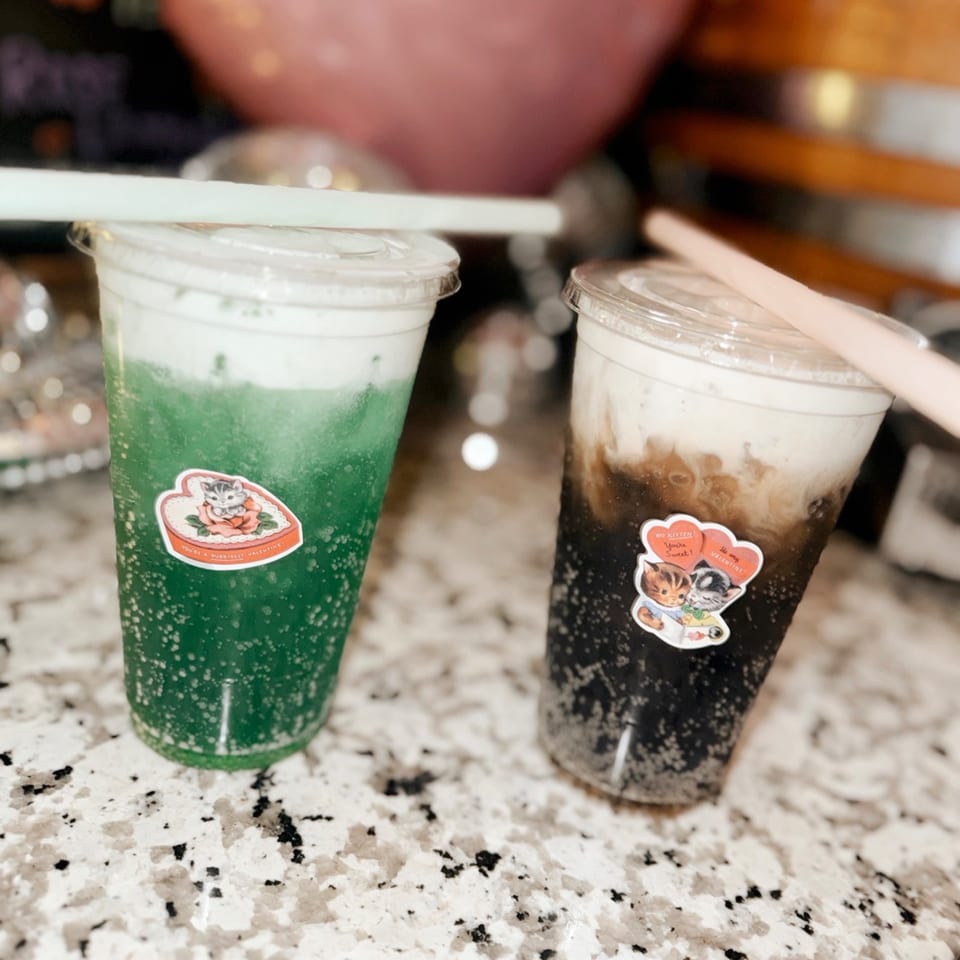 Two drinks with cat-themed Valentine's Day stickers, one green and one dark, on a speckled counter.