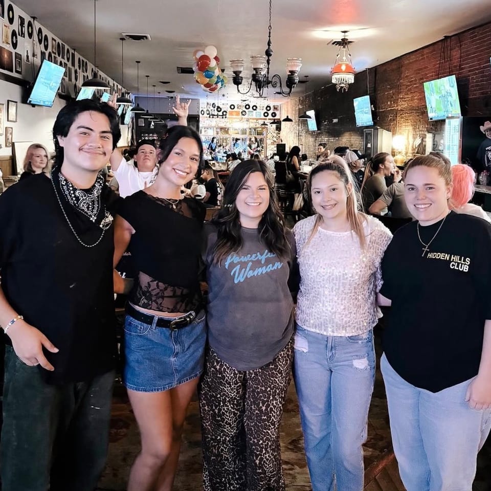 Five young adults smiling at the camera in a lively bar with brick walls and records.