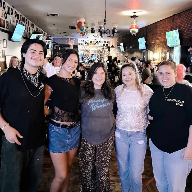 Five young adults smiling at the camera in a lively bar with brick walls and records.