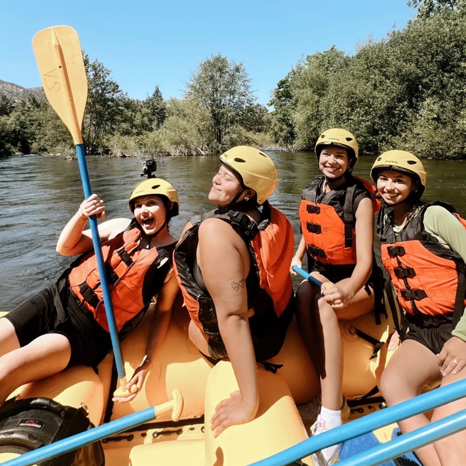 Four women in yellow helmets and orange life vests on a yellow raft, paddling on a river.