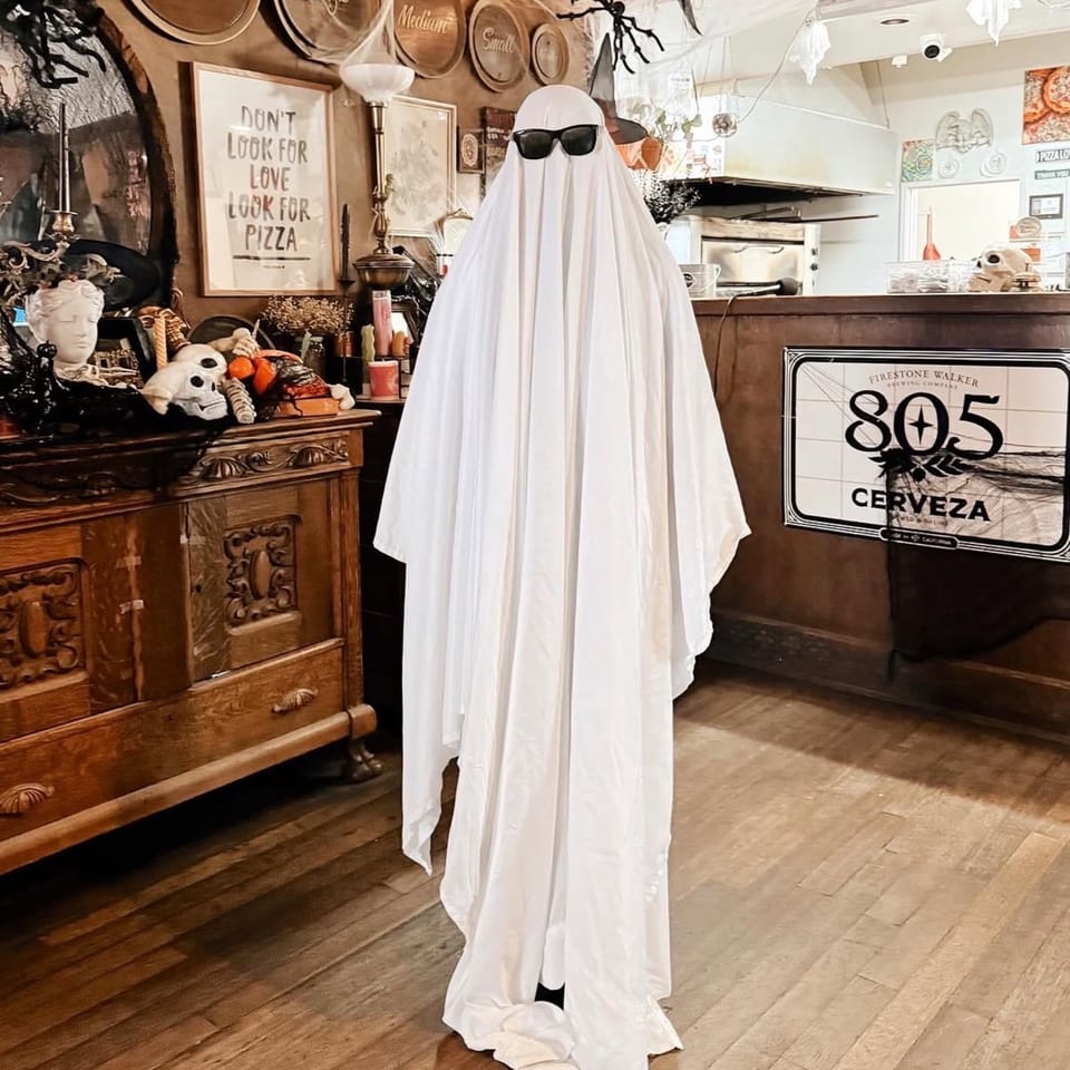 A ghost figure wearing sunglasses stands in a restaurant decorated for Halloween, with a "Look for Pizza" sign.