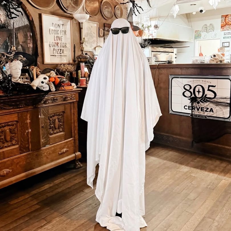 A ghost figure wearing sunglasses stands in a restaurant decorated for Halloween, with a "Look for Pizza" sign.