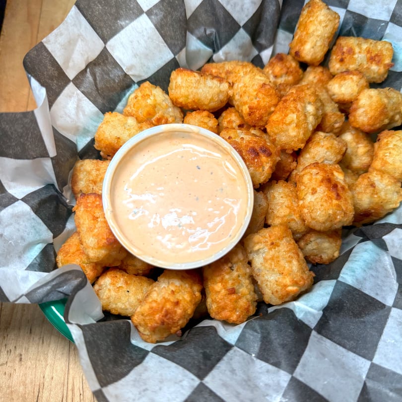 Golden-brown tater tots in a checkered paper-lined basket with a side of dipping sauce.