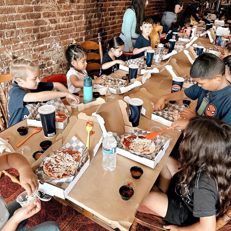 Children at a long table making individual pizzas in a rustic restaurant setting.